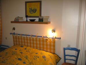 A bedroom with a bed, a chair, a shelf with books, and a wall-mounted light.