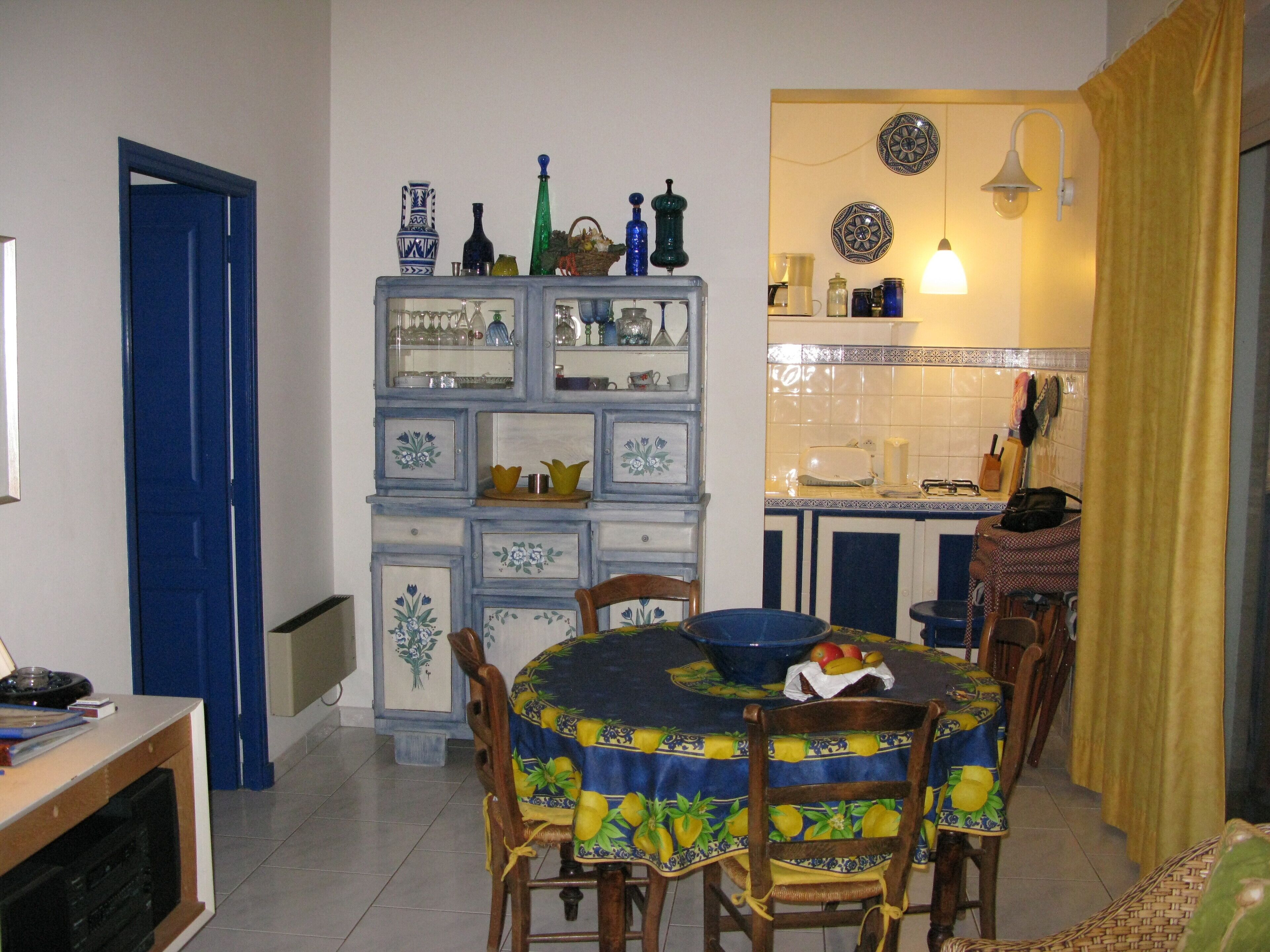 A dining area with a table set for four, surrounded by chairs. A blue cabinet with decorative items is to the left.