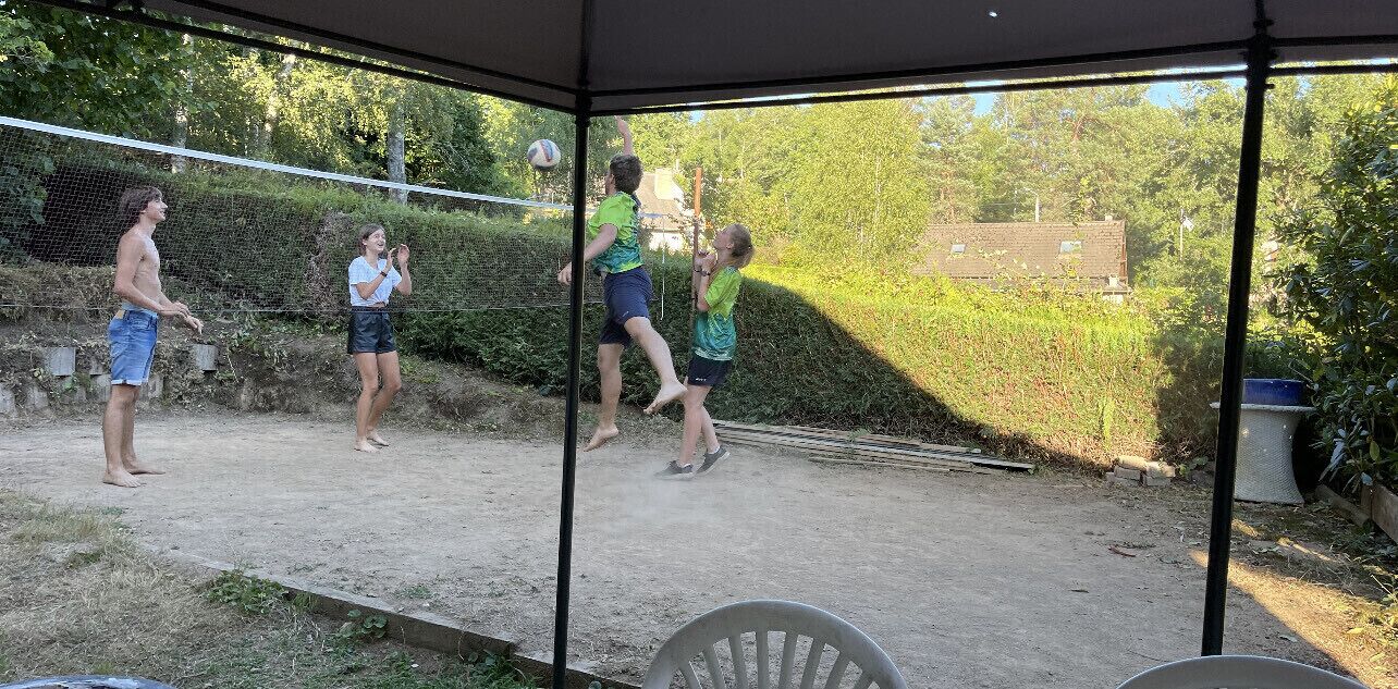 Un espacio al aire libre con una zona de estar cubierta, una red de voleibol y suelo de tierra.