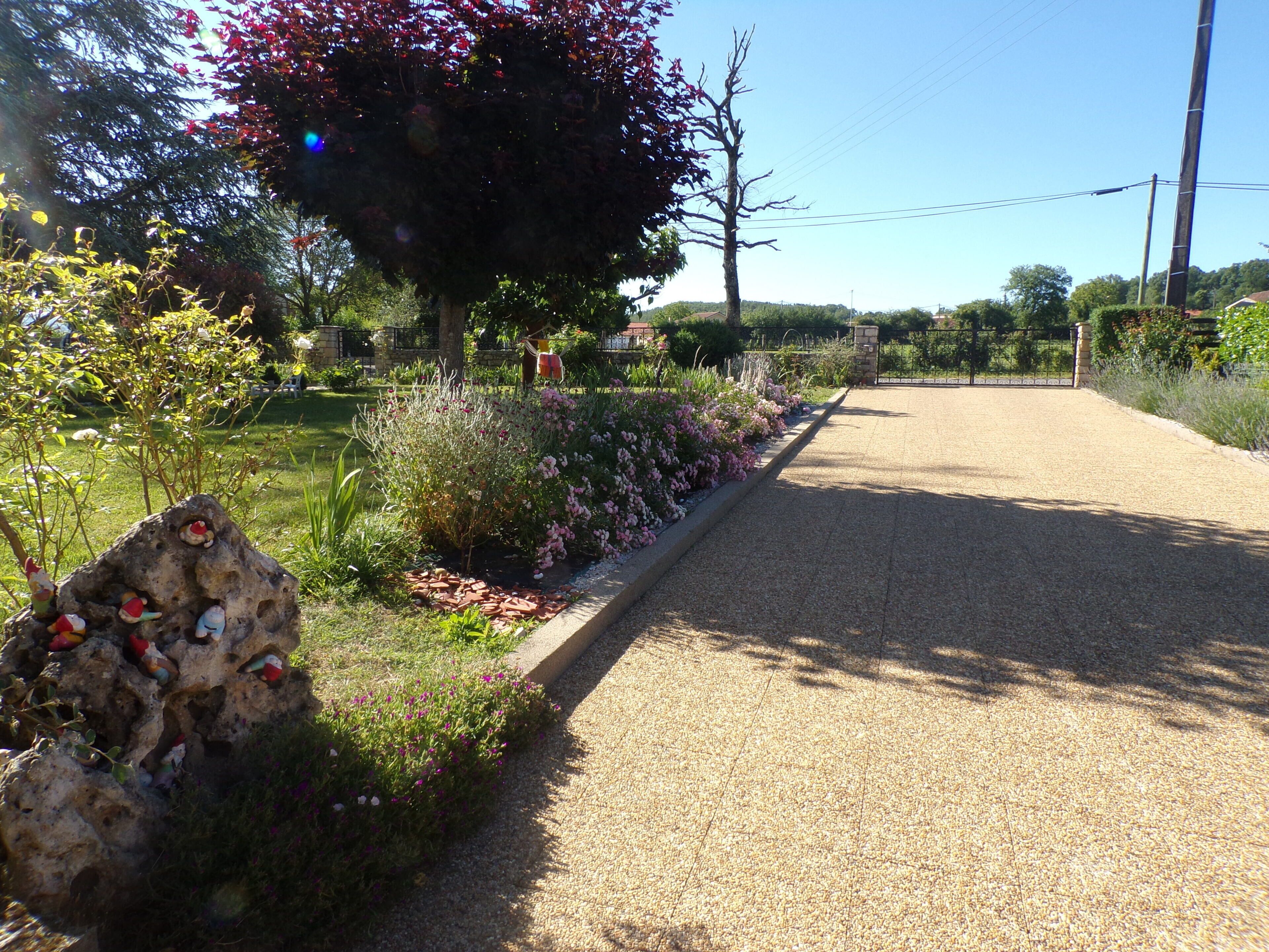 A gravel driveway leading to a gated entrance with a variety of plants and a large tree.