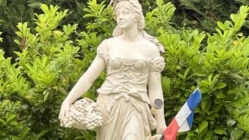 A statue of a woman holding a bunch of grapes and a flag, with a bird perched on her head, set against a backdrop of green foliage.