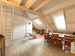A spacious loft with high ceilings, exposed wooden beams, a dining area with wooden chairs and a table, and a cozy seating area with cushions. - Place de la mairie T2 type loft 70m2 terrasse 35m2 (Cauterets)
