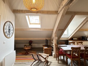 A spacious loft with a high ceiling, exposed wooden beams, a dining area with a long table and chairs, a kitchen area, and a large clock on the wall.
