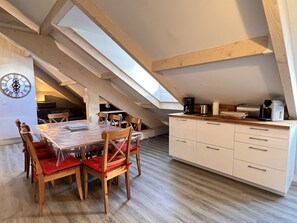 Un espace repas avec une table et des chaises en bois, une cuisine avec des meubles blancs et un plafond en pente agrémenté de poutres en bois.