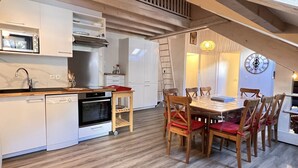 A kitchen with white cabinets, a dishwasher, an oven, and a wooden dining table with chairs. - Place de la mairie T2 type loft 70m2 terrasse 35m2 (Cauterets)