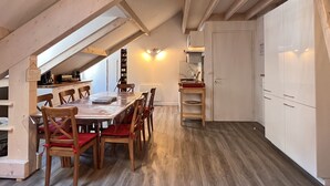 A dining area with a wooden table and chairs, a kitchen with white cabinets, and a sloped ceiling with wooden beams. - Place de la mairie T2 type loft 70m2 terrasse 35m2 (Cauterets)