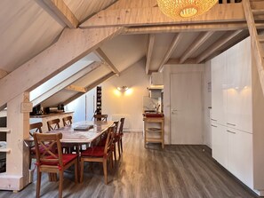 Ein geräumiges Dachzimmer mit hohen Decken, Holzbalken, einem Esstisch mit Stühlen, einer Küchenzeile mit Spüle und Schränken sowie einem großen Kleiderschrank.