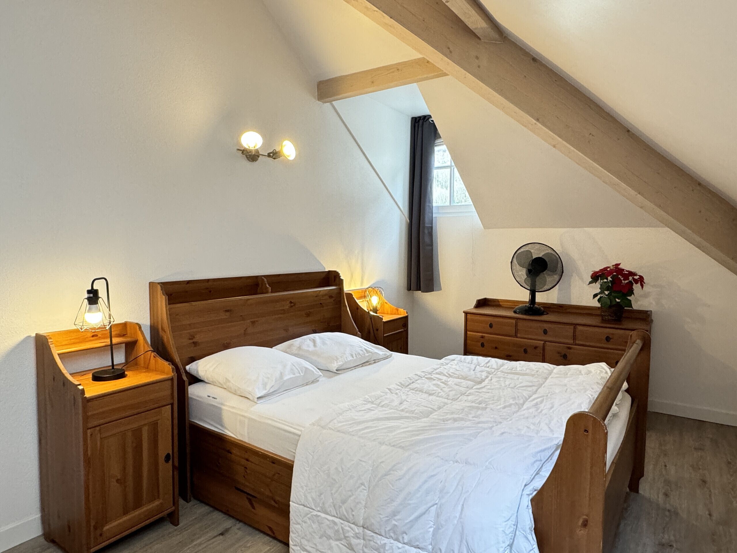 Un dormitorio con techo inclinado, una cama de madera, mesitas de noche, un ventilador de pared y una ventana con cortinas.