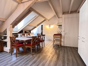 Un espace repas doté d’un plafond haut et d’une verrière, de chaises en bois, d’une table et d’une cuisine avec des meubles blancs.