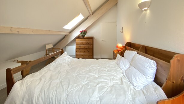 A bedroom with a sloped ceiling, a wooden bed, a nightstand with a lamp, a dresser, and a skylight. - Place de la mairie T2 type loft 70m2 terrasse 35m2 (Cauterets)
