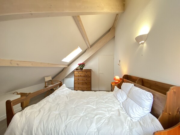 Ein Schlafzimmer mit schrägem Dach, einem hölzernen Bett, einem Schminktisch und einem Nachttisch mit Lampe.
