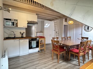 A compact kitchen with a microwave, stove, and dishwasher. There is a dining table with chairs and a ladder against the wall. - Place de la mairie T2 type loft 70m2 terrasse 35m2 (Cauterets)