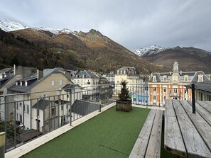 A courtyard with a green lawn, wooden benches, and a view of a town with historic buildings and mountains in the background.