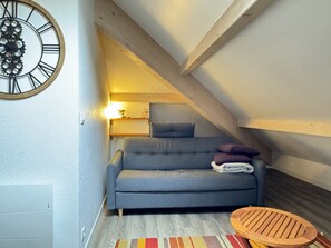 Ein gemütliches Schlafzimmer im Dachgeschoss mit grauem Sofa, einem Holzbalken an der Decke und einer großen Wanduhr.