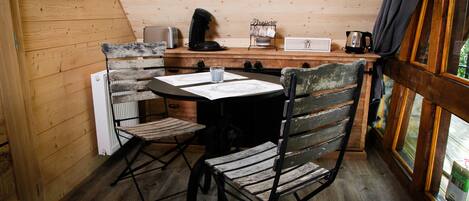 A wooden cabin interior with a dining table, two chairs, a coffee maker, a toaster, and a window with curtains.