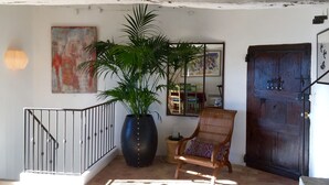 Une pièce avec un escalier, une grande plante en pot, une chaise en bois, une porte en bois et un mur orné d’un tableau.