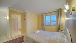 A bedroom with a bed, a window with a city view, a door, and a wall-mounted light fixture. - DUPLEX EXCEPTIONNEL AVEC VUE MER A1B059 (Cannes)