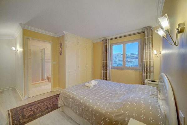 A bedroom with a bed, a window with a city view, a door, and a wall-mounted light fixture. - DUPLEX EXCEPTIONNEL AVEC VUE MER A1B059 (Cannes)
