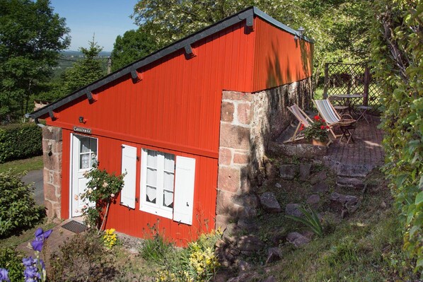 A red building with a stone foundation, a white door, and a small garden area with a bench and a table. - Cassiopée, for 2 to 4 people with exceptional panoramic views (NOAILHAC)