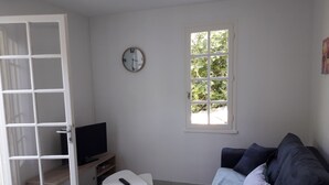 Un soggiorno con una TV su un mobile, un tavolo bianco con un telecomando, un divano scuro, una finestra con vista su del verde e una sveglia a parete.