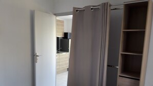 Una camera da letto con un materasso bianco, un armadio in legno e una zona cucina visibile attraverso una porta aperta.