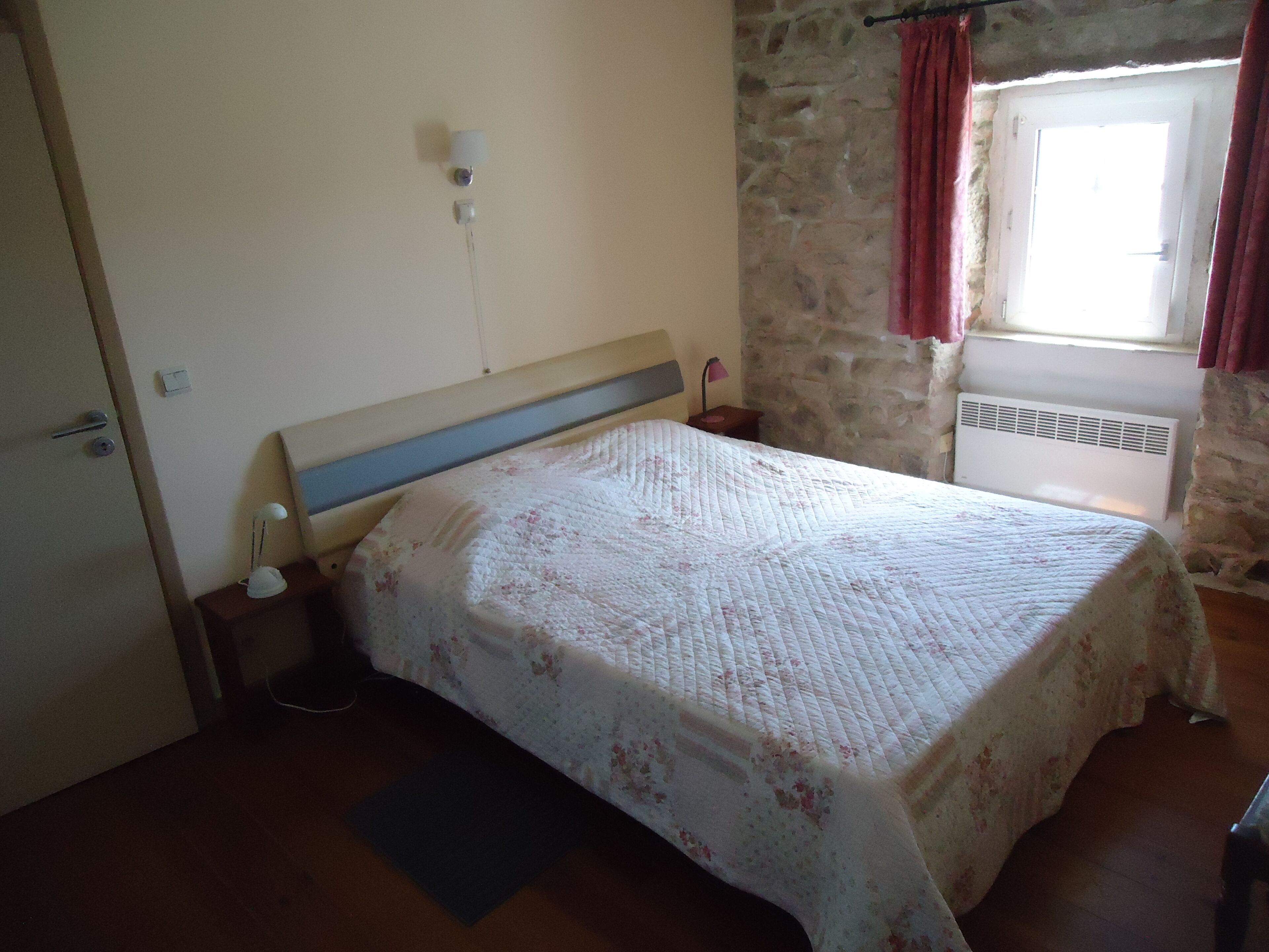 A bedroom with a bed, a nightstand, a wall socket, a window with curtains, and a stone wall.