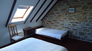 A small, cozy room with a sloped ceiling, a stone wall, a single bed, a chair, and a small table.