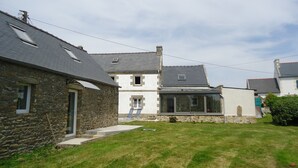 A two-story stone house with a sloped roof, a small garden, and a clear sky.