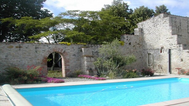 A swimming pool with a stone wall and archway, surrounded by various plants and flowers. - Country house Bedroom B (Lasgraisses)
