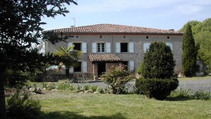 A two-story stone building with a tiled roof, a small front porch, and a well-maintained garden with various plants and a large tree. - Country house Bedroom B (Lasgraisses)