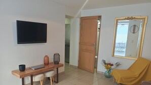 A modern living room with a wooden coffee table, a TV mounted on the wall, a dining area with a wooden table and chairs, and a large mirror with a gold frame.