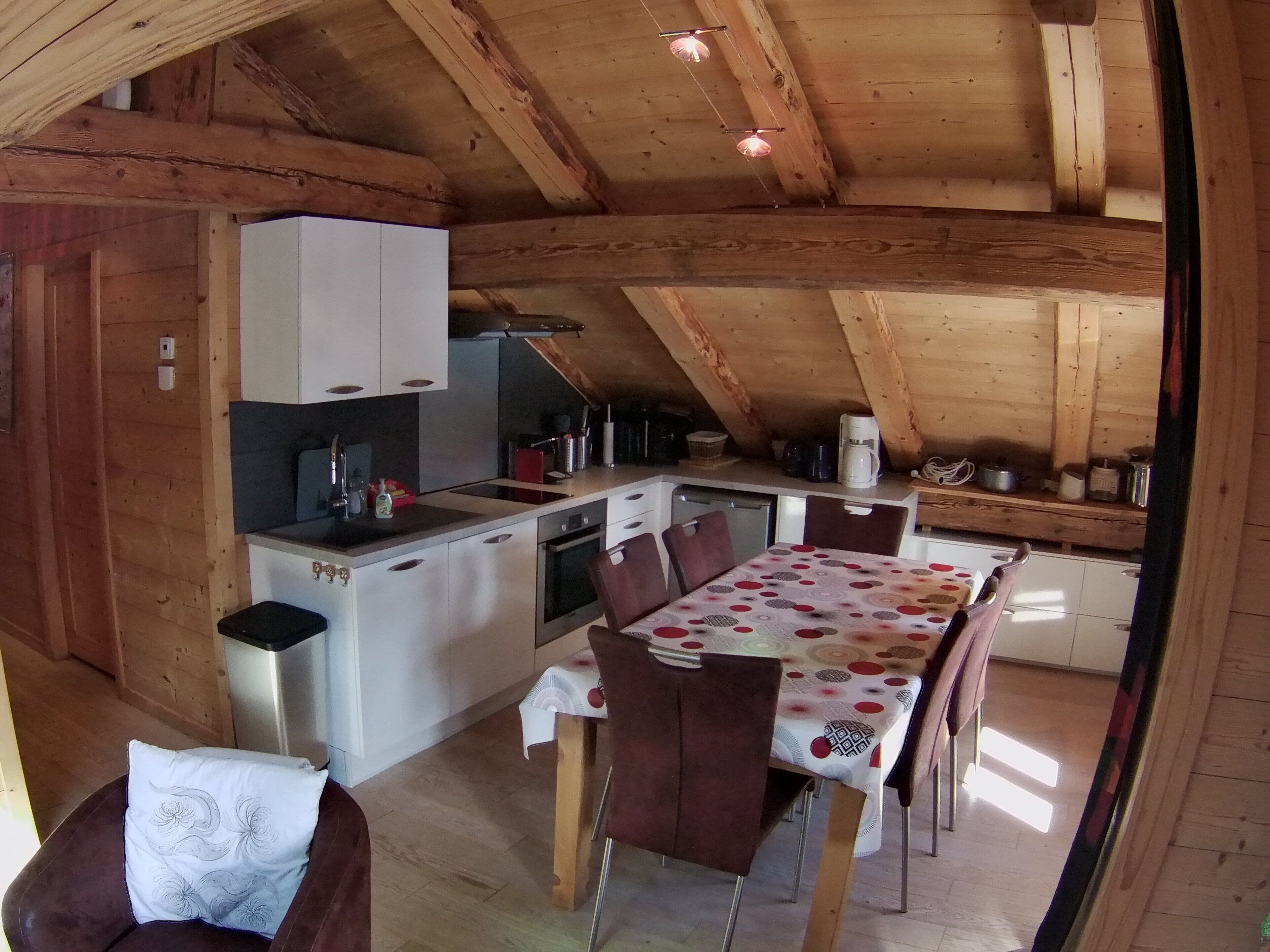 A cozy kitchen with wooden beams, a dining table with chairs, and a small seating area with a cushion.