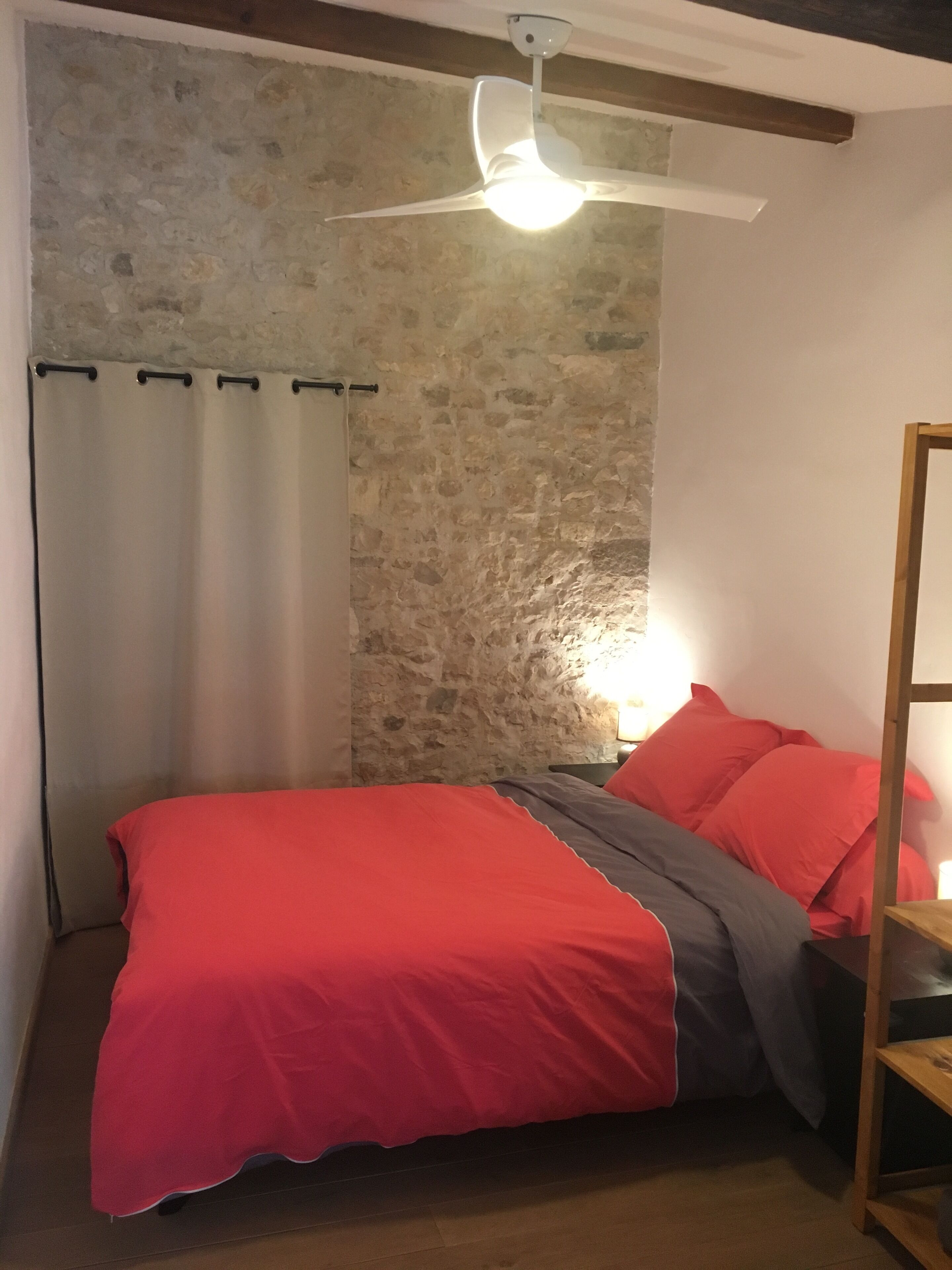 Ein Schlafzimmer mit Steinwand, einem Bett mit rot-grauer Bettwäsche, einem Deckenventilator und einem Holzbodenregal.