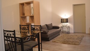 A living room with a sofa, a glass table with chairs, a wooden bookshelf, a lamp, and a rug.