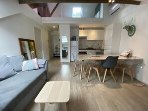A modern living room with a grey sofa, a small wooden coffee table, and a dining area with a glass table and chairs. A kitchen is visible in the background with white cabinets and a tiled backsplash.
