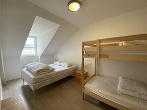 A dormitory room with two bunk beds, a small bedside table, a lamp, and a window providing natural light.