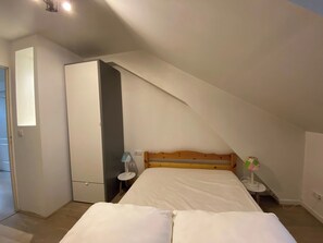 A bedroom with a sloped ceiling, a bed with white bedding, a wooden headboard, a white wardrobe, and a small bedside table with a lamp.
