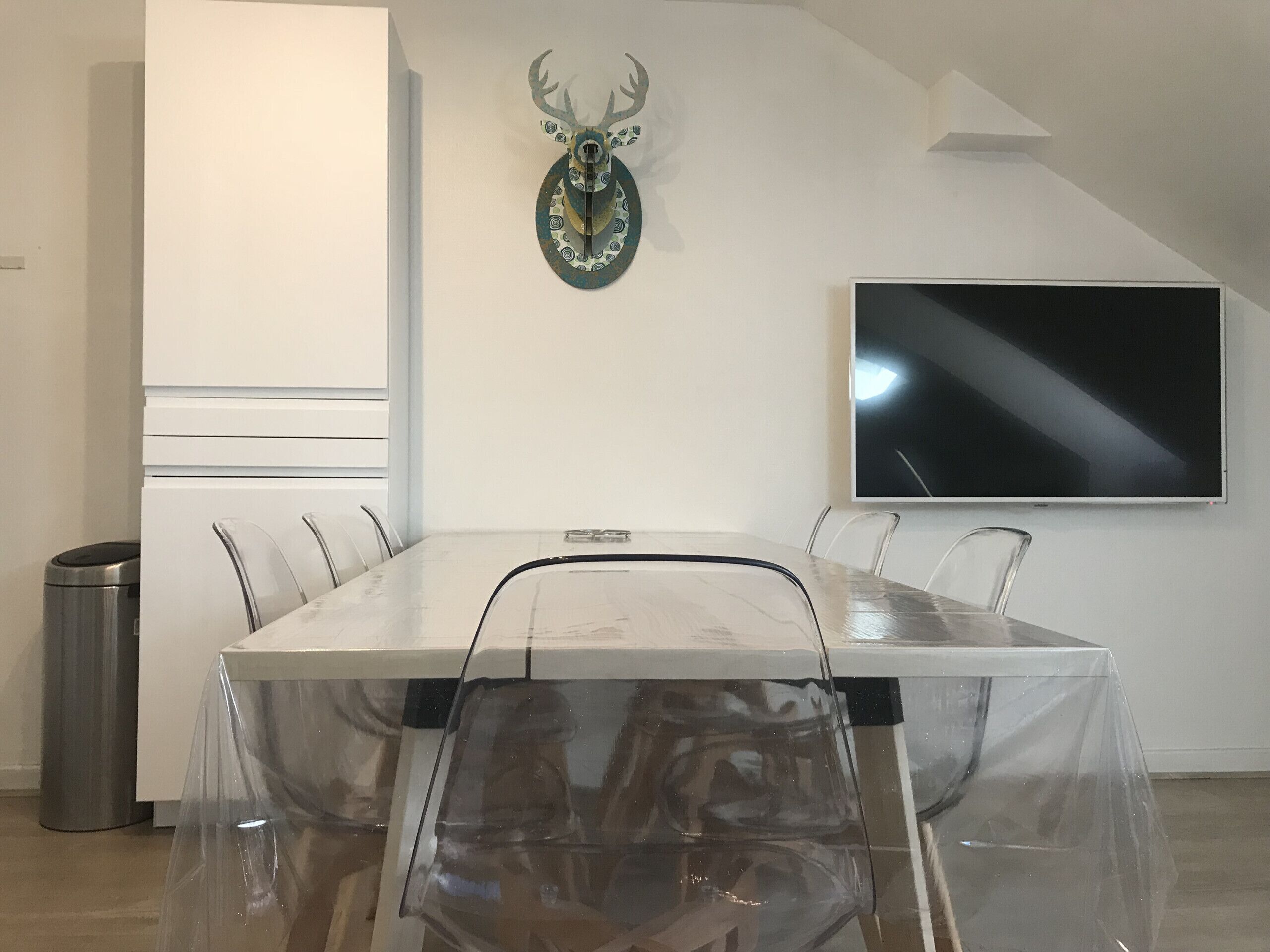 A conference room with a clear table, white walls, a mounted deer head decoration, and a flat-screen TV.