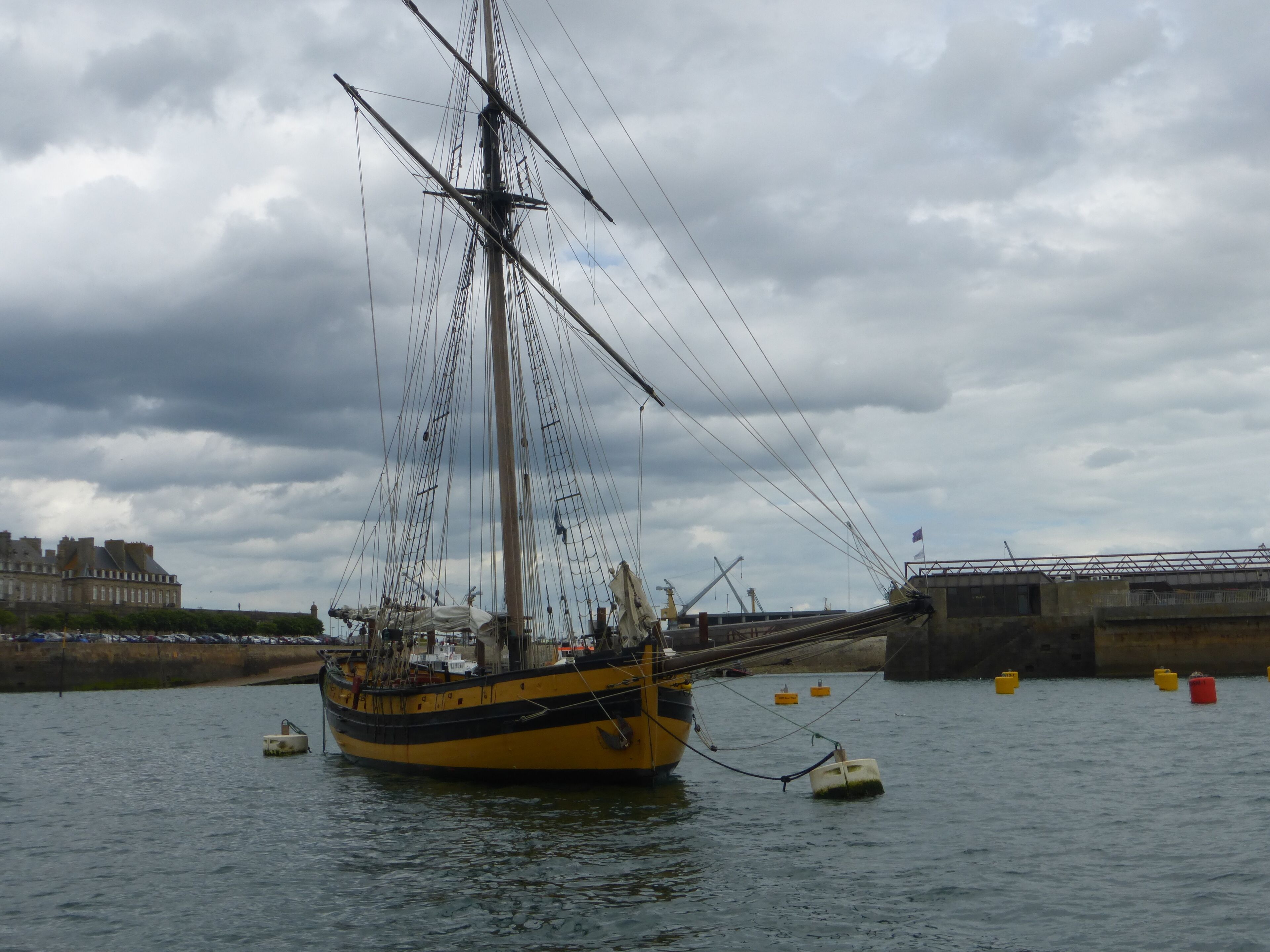 A large sailing ship docked at a harbor with a cloudy sky in the background.