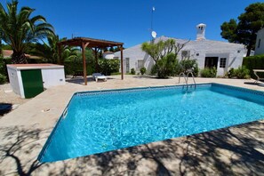A rectangular swimming pool with clear blue water, surrounded by a patio area with a pergola, lounge chairs, and a small building with a green door. - Detached single storey house with garden and private pool Wifi (Ametlla De Mar, L')