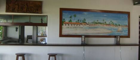 A room with a large beach scene painting, a bar area with stools, and a bed with a patterned bedspread.