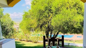 Une table en bois, sur laquelle sont disposés un plateau avec des tasses et une théière, est placée sur une terrasse carrelée, offrant une vue sur un jardin luxuriant et une piscine.