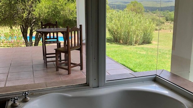 Une salle de bain avec une grande baignoire, une fenêtre donnant sur un jardin et des collines, ainsi qu’une table en bois avec des chaises à l’extérieur.