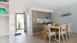 A modern kitchen with white cabinets, a dining area with a wooden table and chairs, and a view of the outdoors through a glass door.