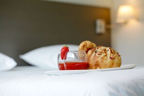 Ein Frühstückstisch mit einem Croissant und einem Glas roter Flüssigkeit auf einem weißen Teller, platziert auf einem Bett mit weißer Bettwäsche und einem Kopfteil im Hintergrund.
