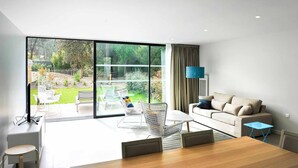 A modern living room with a large sliding glass door leading to a garden, a sofa, a coffee table, and a TV mounted on the wall.