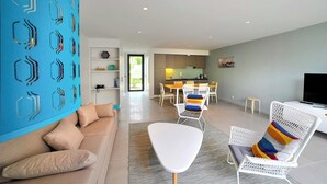 A modern living room with a sofa, a white chair with colorful cushions, a dining area with a table and chairs, and a kitchen in the background.