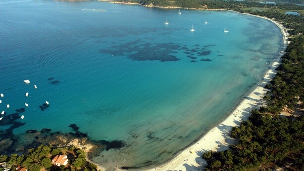 Vista aerea di una zona costiera con spiaggia sabbiosa, imbarcazioni e un faro su una collina.