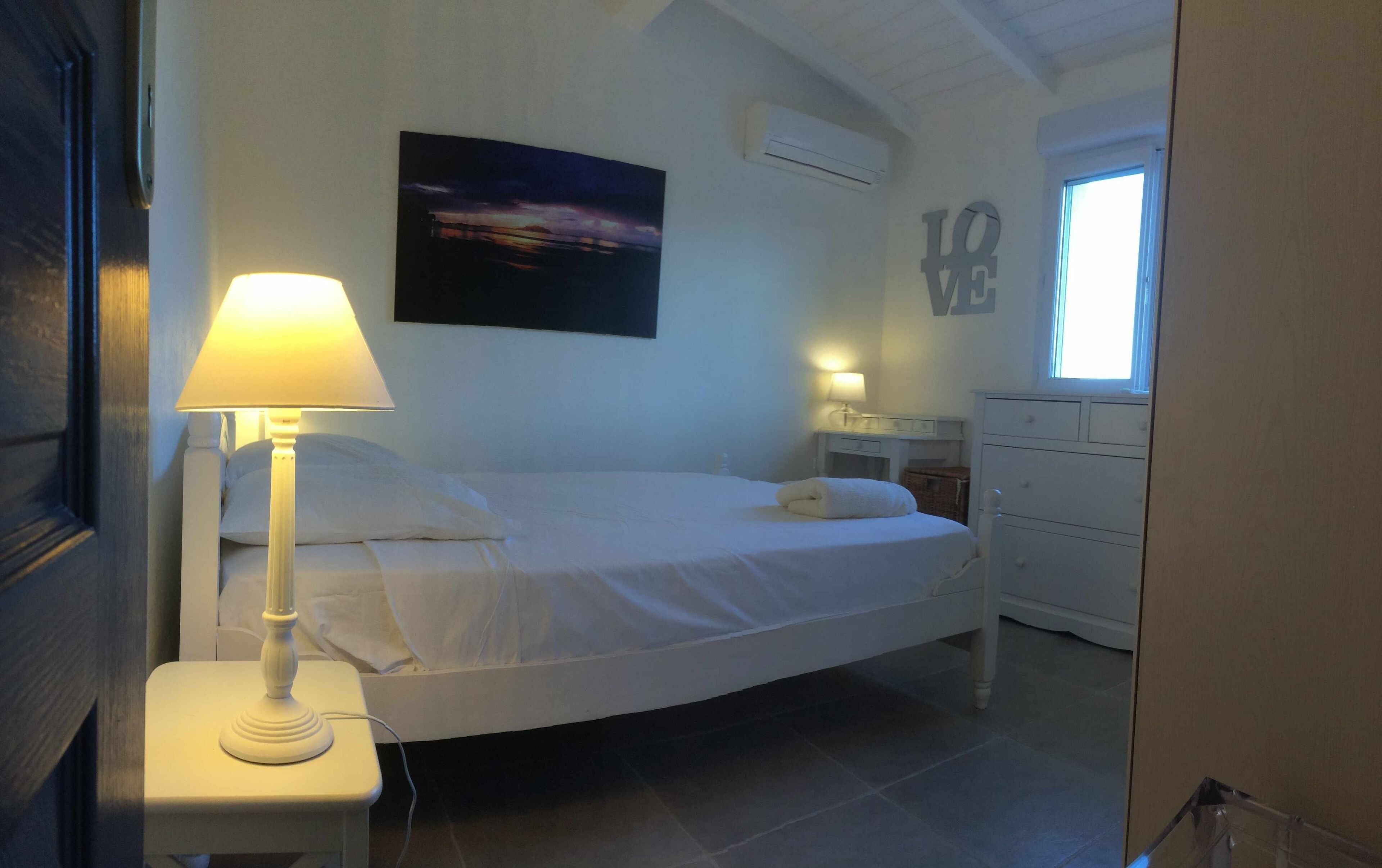 Una cama bien hecha con sábanas blancas, una lámpara de noche, una mesita de noche, un aparador y un cuadro en la pared.