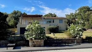 Una casa moderna a due piani con un balcone, immersa nel verde e circondata da un muro in pietra.